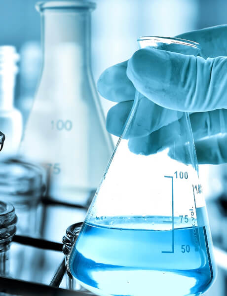 Gloved hand pouring blue liquid into an Erlenmeyer flask in a laboratory.